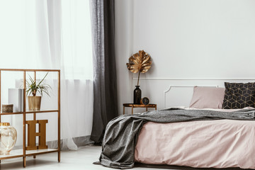 Stylish, golden cabinet and decorations, pastel pink and gray bedding in a bright bedroom interior...