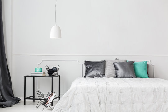 Satin pillows on a minimalist bed by a white empty wall in a trendy bedroom interior with gray and teal green decorations