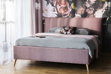 Grey sheets on pink bed in bright modern bedroom interior with flowers print on the wall. Real photo