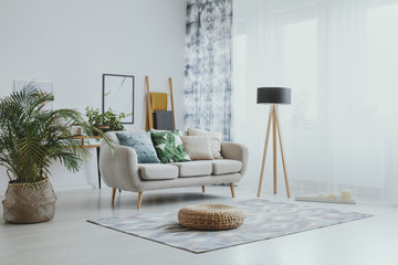 Settee between plant and lamp in bright living room interior with pouf on carpet and poster