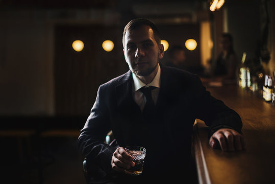 Elegant man in expensive suit near bar counter is smoking cigar and drinking whiskey