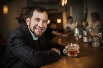 Elegant man in expensive suit near bar counter is smoking cigar and drinking whiskey