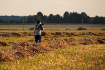 A young white man with a camera walking on the field and taking pictures.