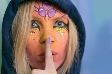 Sorceress, fairy, a woman with a painted face in her hood holds her index finger on her lips.
