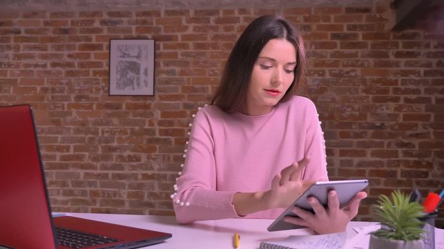 caucasian young businesswoman scrolling tablet concentrated ext to camera on brick background