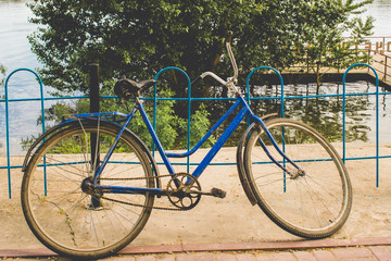 Fototapeta na wymiar Soviet land bike. The simplest transport manual control in vintage style and warm colors. Stands on the background of the pier with green summer trees, a metal fence of blue pipes. 