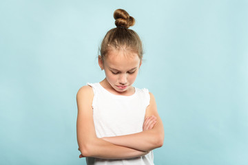 offended grumpy hurt child with fold hands looking down. young cute huffy girl portrait on blue background.