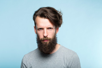 Fototapeta premium serious frowning man with a troubled look and gathered brows. emotion and feelings concept. portrait of grumpy bearded guy on blue background.