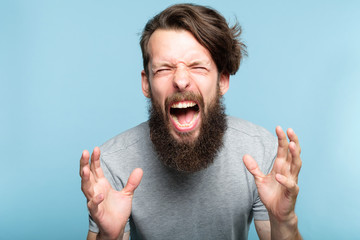 emotional breakdown. angry enraged infuriated crazy man screaming. portrait of a young bearded guy on blue background. emotion facial expression. feelings and people reaction.