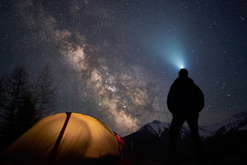 Man tourist with flashlight stands near his camp tent at night under a sky full of stars.