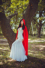 Obraz na płótnie Canvas Halloween concept, glamorous costume detail. Young beautiful and mysterious woman in woods, in white Dress and Red Cloak. Little red hood or vampire story
