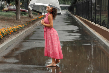 Young girl in a red dress with a transparent umbrella dancing in the rain standing in a puddle