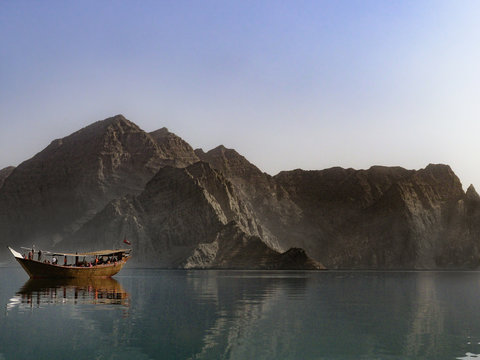 A traditional dhow boat sits in front of rocky mountains in the calm waters of Khor Ash Sham in the fjords of the Musandam Peninsula in northern Oman