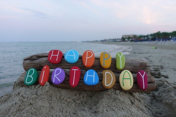 Happy Birthday message with multi colored stones over two pieces of wood with sea background