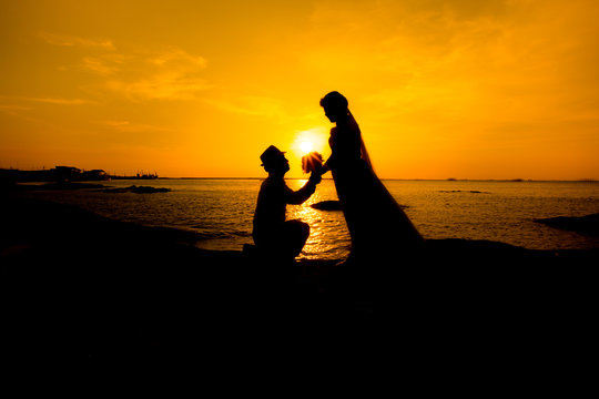 A silhouette of a young man, down on one knee and holding a bouquet, proposing to his girlfriend.will you marry me images.Young couple in love at beautiful sunset.inspired by glistening gold.