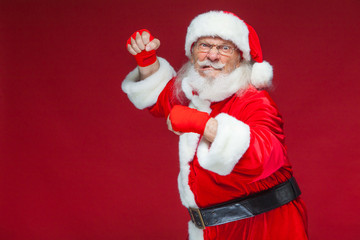 Christmas. Santa Claus with red bandages wound on his hands for boxing imitates kicks. Kickboxing, karate, boxing. Isolated on red background.