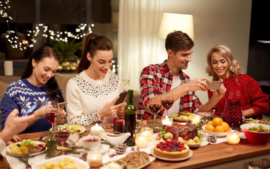 holidays and celebration concept - happy friends with smartphones having christmas dinner at home