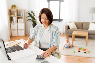 multi-tasking, freelance and motherhood concept - working mother with laptop computer counting on calculator and baby boy playing at home office