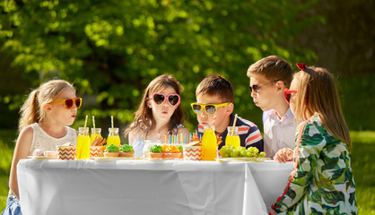 holidays, childhood and celebration concept - happy kids blowing out candles on birthday cake sitting at table at summer garden party