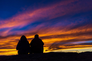 A man and a woman are looking at a beautiful sunset. They sit on a hill. Silhouette