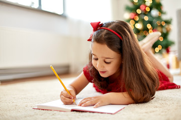 christmas, holidays and childhood concept - smiling girl writing wish list or letter to santa at...