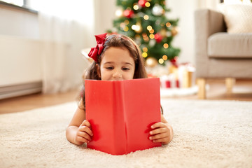 christmas, holidays and childhood concept - happy little girl lying on floor and reading book at...