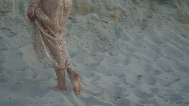 long female legs go along the dune in the sand. the woman gently tramples the sand. a long golden dress develops in the wind. 4K