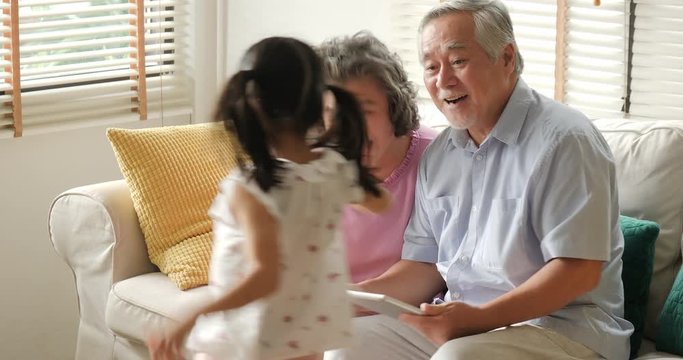 Little girl comes to hug her grandfather and grandfather while they playing tablet. Kid playing with senior man and woman with happy emotion. people with family, lifestyle, technology concept.