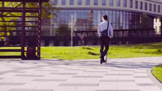 Dolly shot. A man in a business suit riding a kick scooter in a park then he taking scooter and going up stairs. Rear view