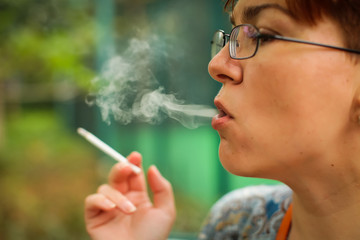 Fototapeta premium woman smokes thin cigarette, exhaling smoke. Female hand with smoking cigarette on blurred green park background