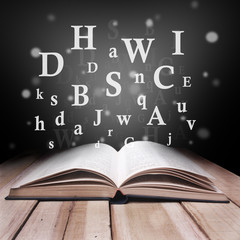 Opened Book on Wooden Table Against Black Dark Background with Letters Flying