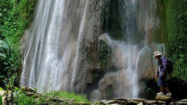 Senior man Asian enjoying walking in front of the waterfall. Travel Lifestyle and success concept vacations into the wild wearing cozy shirt,hat with backpack. Video Slow motion