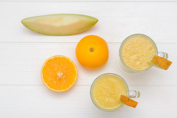 Two melon smoothie cups, a cut orange and a piece of melon on a white table. The view from the top. Flat lay.