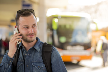 Portrait of young handsome smiling man waiting at the bus stop with modern designed smartphone in hand. Traveling by eco friendly alternative to car. Nice summer sunny weather and warm colors photo.