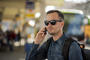 Portrait of young handsome smiling man waiting at the bus stop with modern designed smartphone in hand. Traveling by eco friendly alternative to car. Nice summer sunny weather and warm colors photo.
