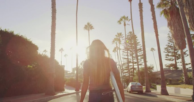 Attractive surfer girl walking with skateboard down street at sunset with lens flare 
