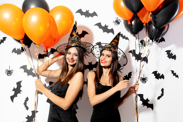Two charming young women in witch's hats hold black and orange balloons on a white background with...