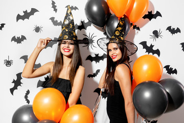 Two beautiful young women in witches hats are with black and orange balloons on a white background...