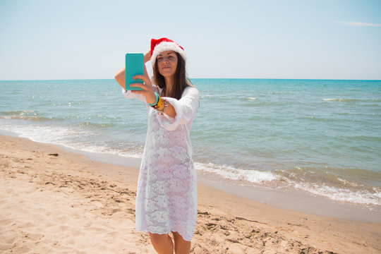 Christmas young smiling woman in red santa hat taking picture self portrait on smartphone at beach over sea background. toned