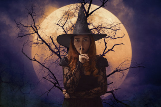 Halloween witch showing silence sign with finger over lips standing over dead tree, full moon and spooky cloudy sky, Halloween mystery concept