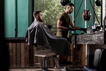 Brutal man with beard sits in a chire at a barber shop. Handsome barber takes the hairclipper