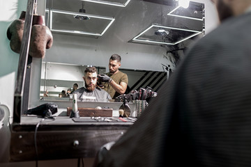 Reflection in the mirror of brutal man sits in a chair and barber shaves his hairs.