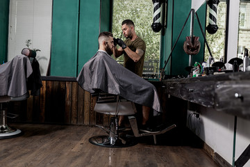 Brutal man with tha beard sits in the chair in front the mirror at a barber shop . Barber shaves mans hairs