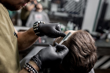 Fototapeta na wymiar Barber in black gloves trims temple of brutal bearded young man with a straight razor at a barbershop