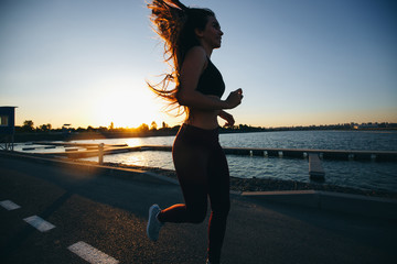 Fototapeta premium Gorgeous young girl with long brown hair dressed in sports clothes runs on the road along the reservoir on the sunset