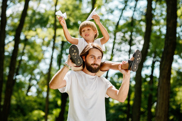 Obraz premium Little boy with his father dressed in the white t-shirts walking in the forest. Boy is sitting on the shoulders of his father and keeps paper airplanes in his hands.