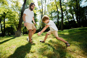 Dark-haired father and his little son dressed in the white t-shirts are playing football on a lawn on a warm day.