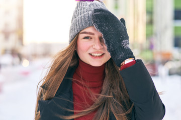 The red-haired young girl in mittens and a cap fervently smiles and corrects a cap on the head. Clothes covered with snow because of playing with friends in the snow. Outdoor recreation in winter