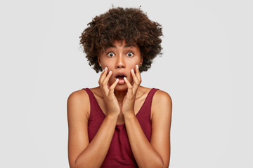 Fototapeta na wymiar Studio shot of shocked African American girl with surprised expression, stares with bugged eyes, keeps mouth opened, dressed in casual red t shirt, poses against white background. Scared black lady