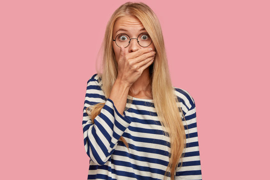 Astonished attractive woman with blonde hair covers mouth, doesnt want to spread rumours, shocked to hear sudden news from interlocutor, dressed in striped casual sweater, isolated on pink wall